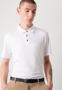 Witte polo shirt met een zachte textuur, drie zwarte knopen en een klein logo op de borst, gecombineerd met beige broeken en een zwarte riem.