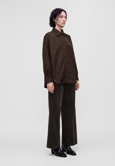 Femme aux cheveux courts et foncés portant une chemise ample marron foncé, un pantalon large assorti et des bottines noires, debout devant un fond uni.
