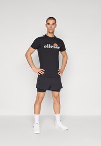 Melnas īsām piedurknēm t-krekls ar "ellesse" logo un sarkanu akcentu, savienots ar melnām sporta biksēm un baltām kedām. Vienkārša fona.