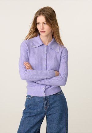 Junge Frau mit langen Haaren, die eine lavendelfarbene Strickjacke mit Zopfmuster und dekoriertem Kragen sowie blaue Jeans trägt, mit verschränkten Armen, schaut zur Seite.