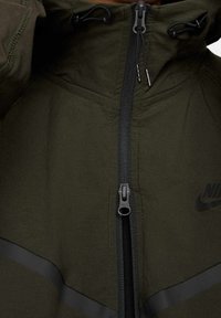 Olivgrön jacka med hög krage, svart dragkedja och en subtil svart logotyp. Har en texturerad tyg och reflexränder.