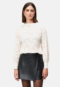Cremiger Strickpullover mit strukturiertem Blumenmuster, kombiniert mit einem schwarzen Leder-Wickelrock mit seitlichem Knopf. Beide Teile sind figurbetont.