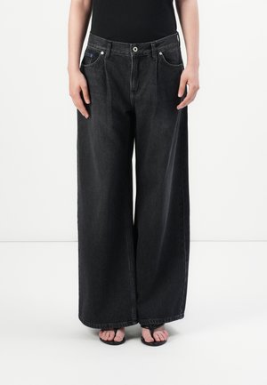 Person iført sorte jeans med vide ben og sorte thong-sandaler foran en ensfarvet hvid baggrund.