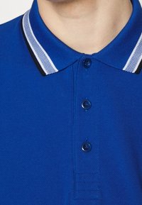 Blå polotröja med texturerad tyg, tre-knappsstängning och krage med svarta och vita randiga detaljer.