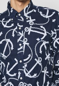 Chemise bleu marine ornée d'un motif de ancres et de cordes blanches. Design à boutons, avec un col classique et des boutons blancs.