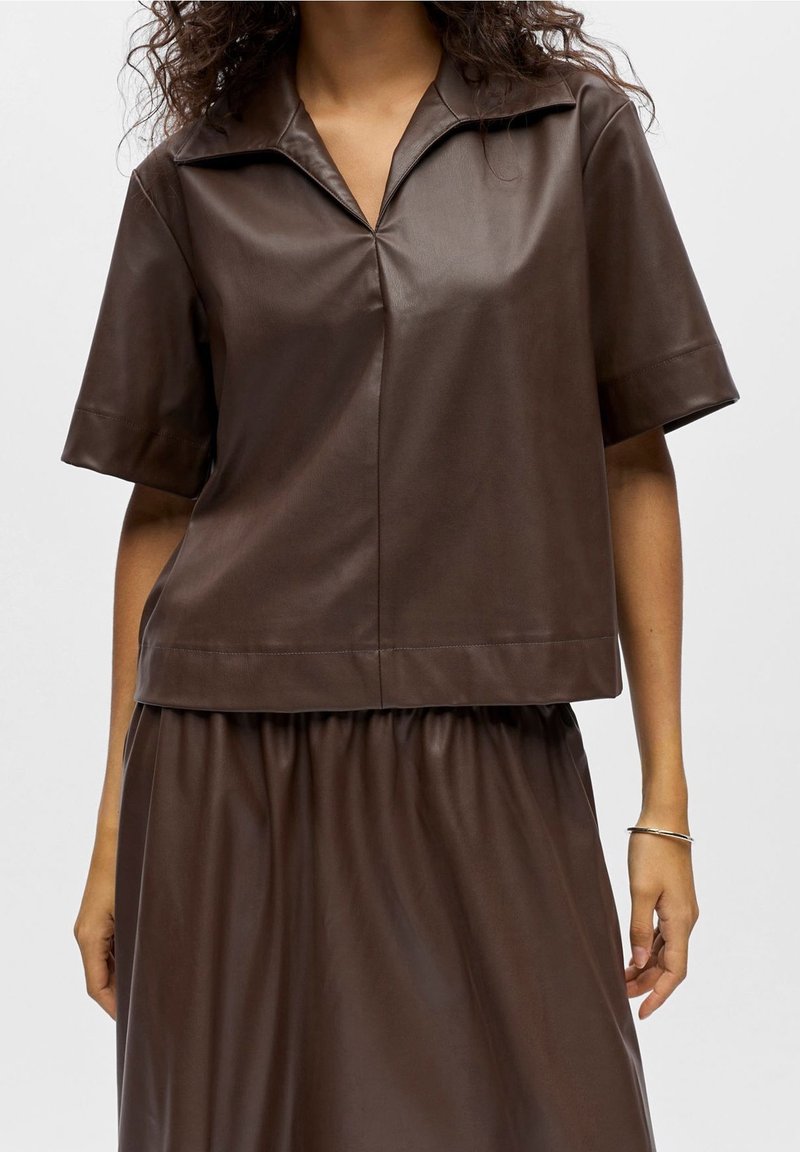 Femme portant une chemise en cuir marron foncé à manches courtes avec col et une jupe en cuir assortie, avec un bracelet en argent à son poignet droit.