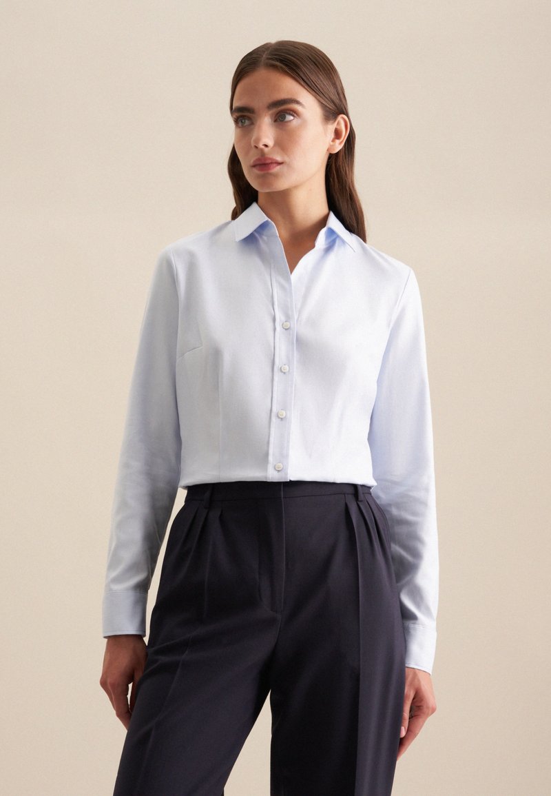 Lichtblauw overhemd met lange mouwen, een puntige kraag en een sluiting met knopen aan de voorkant. De stof is soepel en heeft een enkele borstzak. Gekomplementeerd met donkere plooibroek.