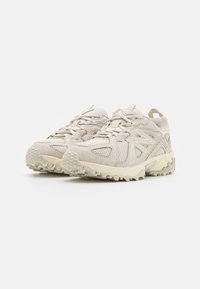 Scarpe da ginnastica beige con materiali in rete e sintetici, dotate di suole con trazione evidente, dettagli testurizzati e sistema di lacci rinforzato.