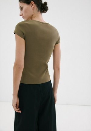 Donna con capelli scuri raccolti in uno chignon, indossa una maglietta aderente a maniche corte color verde oliva e pantaloni neri larghi, in piedi davanti a uno sfondo bianco semplice.