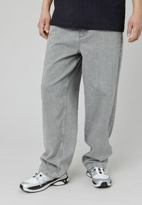 Pantalons en denim gris clair avec une coupe ample, des coutures contrastées et des poches latérales, associés à des baskets noir et blanc.
