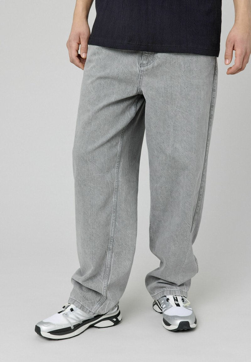 Pantalons en denim gris clair avec une coupe ample, des coutures contrastées et des poches latérales, associés à des baskets noir et blanc.