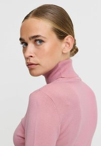 Šviesiai rožinė, prigludusi turtleneck tipo megztinis su ribotu tekstūru. Aukštas apykaklė ir ilgomis rankovėmis, minimalūs siūlai peties ir nugaros srityse.