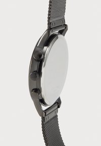 Montre noire en métal avec un boîtier rond, un bracelet en maille et deux boutons en haut. L'arrière est lisse et réfléchissant, marqué des détails de la marque.