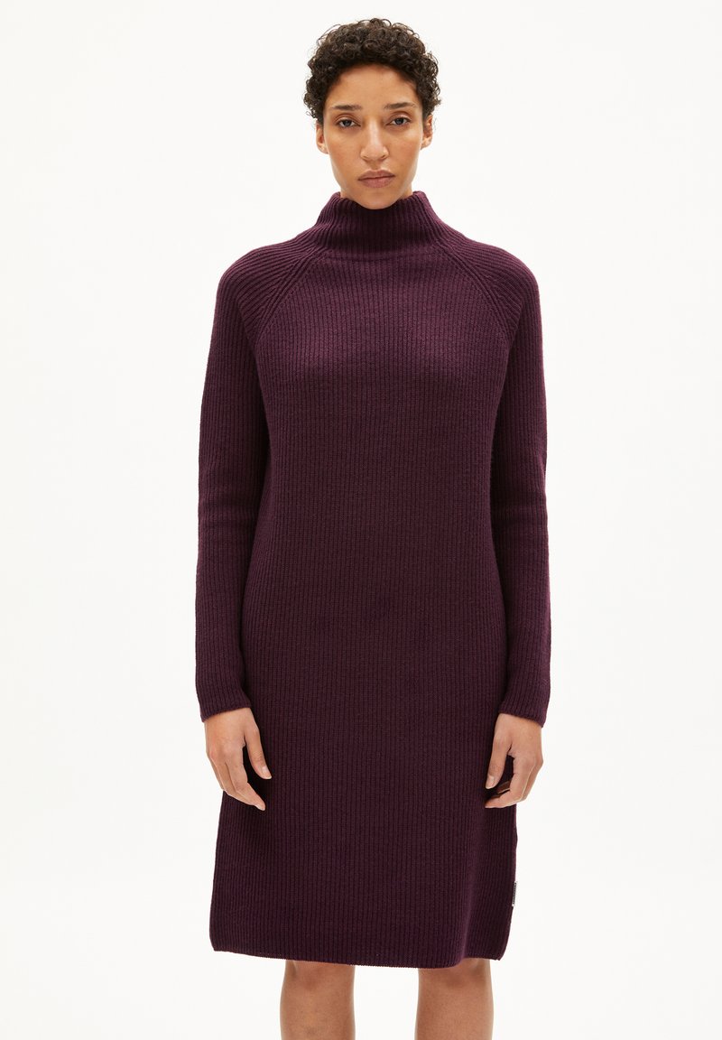 Robe en tricot de couleur violet profond avec un col montant, des manches longues et une texture côtelée ; longueur genoux avec un détail de fente sur le côté.