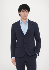 Sötétkék zakó keskeny hajtókákkal, két gombbal és elülső zsebekkel, világoskék inggel párosítva. Arany kitűző a hajtókán.