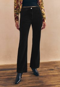 Pantaloni din catifea neagră, evazați, cu talie ajustată și buzunare frontale, asortați cu cizme cu vârf albastru și o cardigan cu motive.