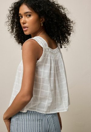 Žena s kučeravými vlasmi, oblečená v bielom tielku bez rukávov s čipkovaným detailom a modrých pruhovaných nohaviciach, pozerá cez rameno.