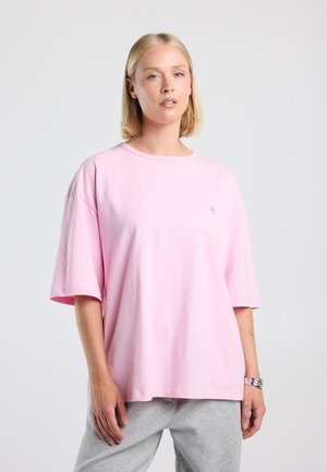 Jonge vrouw met kort blond haar, die een loszittend lichtroze T-shirt en grijze broek draagt, staand tegen een eenvoudige lichte achtergrond.