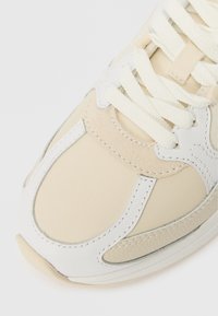 Κοντινό πλάνο της μύτης ενός μπεζ και λευκού αθλητικού παπουτσιού με υφή δέρματος, πάνελ σουέτ και χοντρές λευκές κορδόνες σε λευκό φόντο.