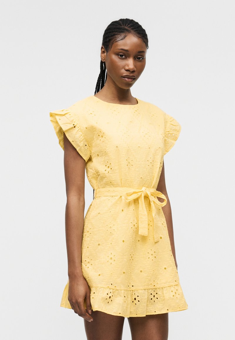 Γυναίκα που φορά κίτρινο φόρεμα με τρυπητά σχέδια, με φουσκωτά μανίκια, δεμένη ζώνη στη μέση και ραμμένο βολάν στο τελείωμα, στέκεται μπροστά σε ένα απλό φωτεινό φόντο.