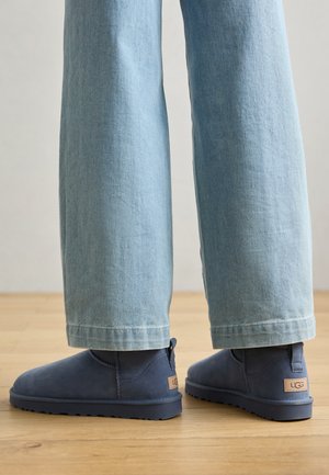 Stivaletti alla caviglia in suede blu navy con suola piatta, dotati di una linguetta posteriore e di una piccola toppa con logo. Indossati con jeans a gamba larga di colore blu chiaro.