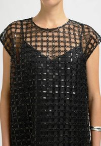 Moteris, apsirengusi juodu blizgučiais išmargintu tinklu, po juodu marškinėliais, su sidabriniu apyrankiu ant dešinės rankos, prieš vientisą foną.