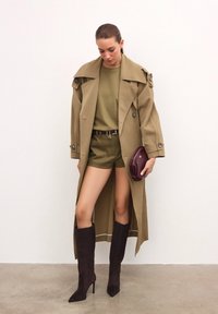 Bežki trench kaput preko maslinasto zelene majice i kratkih hlača, uparen s crnim čizmama do koljena i bordo clutch torbicom, prikazan na neutralnoj pozadini.