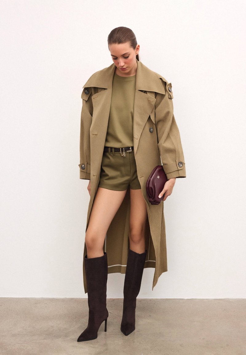 Bežki trench kaput preko maslinasto zelene majice i kratkih hlača, uparen s crnim čizmama do koljena i bordo clutch torbicom, prikazan na neutralnoj pozadini.