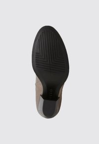 Semelle en caoutchouc noir avec un motif de semelle géométrique et un talon carré sur une chaussure en cuir beige, présentée par le dessous sur un fond uni.