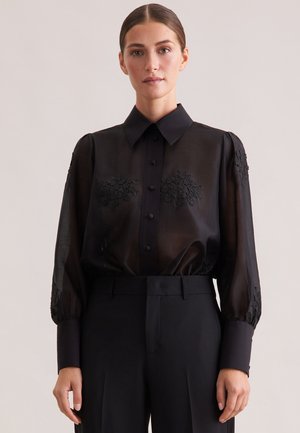 Kvindelig iført en gennemsigtig sort bluse med blomsterbroderi, knapmanchetter og bred krave, stoppet ned i høj taljede sorte bukser.