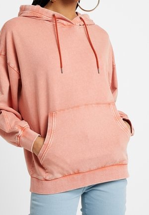 Vrouw draagt een oversized vervaagde koraalkleurige hoodie met een voorzak en trekkoorden, handen in de zakken gestoken, gecombineerd met lichtblauwe spijkerbroek.