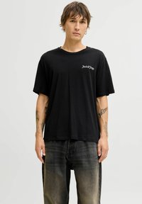 Camiseta negra de corte relajado, con mangas cortas y un logo blanco en el lado izquierdo del pecho. Combinada con unos vaqueros negros desgastados.