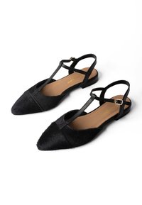 Melni punkti pēdas kurpju ar T-veida siksnu un potītes sprādzi, ar teksturētu augšdaļu un brūnu polsterētu zolīti, attēloti uz balta fona.