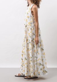 En kvinde modellerer en lang, ærmeløs hvid kjole med gule og grønne blomster mønstre, parret med grå sandaler, mod en neutral baggrund.