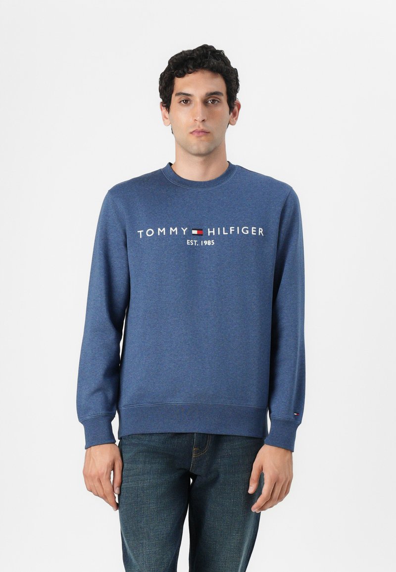 Mėlyna medvilninė džemperis su apvaliu kaklu, nertomis rankovėmis ir apačia; priekyje yra "TOMMY HILFIGER" logotipas ir įsteigimo datos grafika.