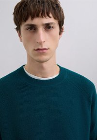 Suéter de punto color verde azulado con un patrón acanalado texturizado y escote redondo, superpuesto sobre una camiseta blanca de cuello redondo. Diseño sencillo y ajustado.