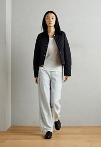 Veste matelassée noire avec quincaillerie argentée, chemise blanche, jean large bleu clair, et chaussures noires ; modèle marchant sur un sol en bois contre un mur gris.