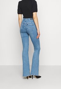 Frau, die mit dem Rücken zur Kamera steht und hellblaue hochgriffige Schlagjeans, ein schwarzes kurzärmeliges Oberteil und schwarze spitze High Heels trägt, vor einem einfarbigen Hintergrund.