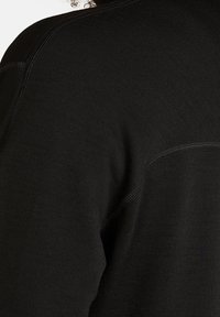 Nærbillede af sort stof med synlige sømme langs skulder- og ærmesømme, der fremhæver tekstur og detaljer i beklædningens konstruktion.