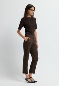 Žena v tmavě hnědém topu s krátkým rukávem a ozdobami, elegantních kalhotách stejné barvy, černých lodičkách s T-páskem a brýlích, stojí s rukou v kapse.