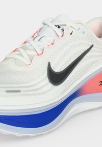 Λευκό αθλητικό παπούτσι Nike με μαύρο λογότυπο, λευκά κορδόνια και επενδυμένη σόλα με μπλε και ροζ διαβαθμισμένες λεπτομέρειες.