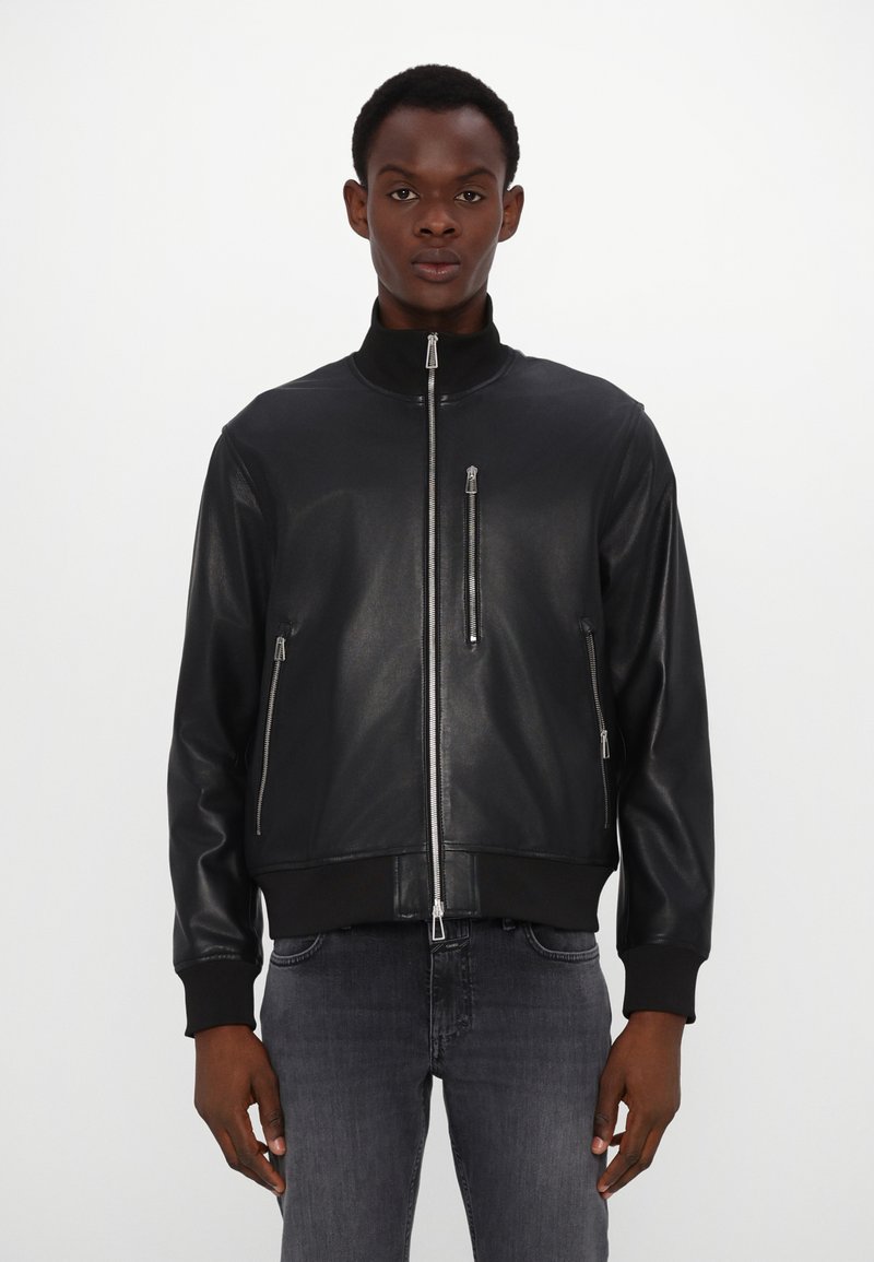 Νεαρός άνδρας που φορά μαύρο δερμάτινο μπουφάν με φερμουάρ και ριγωτά μανίκια και γιακά, συνδυασμένο με ξεθωριασμένα μαύρα τζιν, που στέκεται μπροστά σε απλό φόντο.