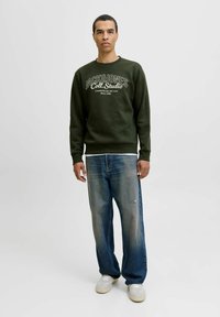 Mand iført en mørkegrøn Jack & Jones sweatshirt, slidte løse blå jeans og lysegrå sneakers, der står foran en hvid baggrund.