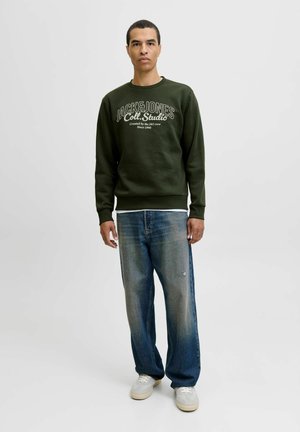 Mężczyzna w ciemnozielonym swetrze marki Jack & Jones, w wyblakłych niebieskich dżinsach o luźnym kroju i jasnoszarych sneakersach, stojący na białym tle.