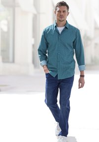 Türkises Button-Up-Hemd mit kleinem Muster, hochgekrempelten Ärmeln, kombiniert mit blauen Jeans und weißen Turnschuhen, die auf einer hellen Oberfläche gehen.