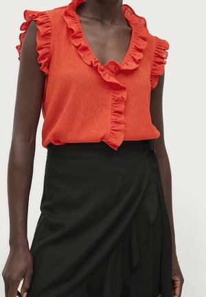 Blouse rouge texturée sans manches avec encolure à volants portée avec une jupe portefeuille noire taille haute avec détail volanté sur le côté.