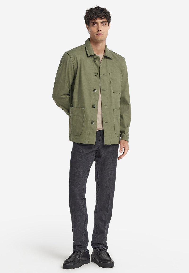 Olivgrön lättviktsjacka med knappfram, två stora framfickor och rak passform. Buren över en beige skjorta och mörka jeans.