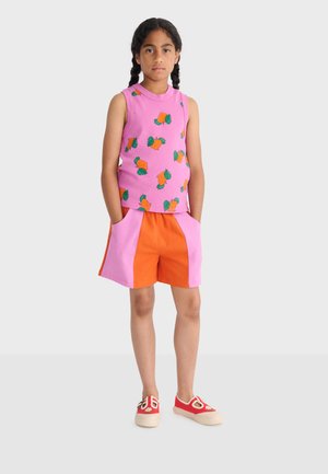 Djevojka s pletenicama, odjevena u ružičastu majicu bez rukava s natpisom mrkve, narančaste i ružičaste kratke hlače i crvene cipele na navlačenje.