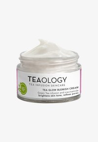 Barattolo di vetro chiaro contenente crema bianca, etichettato "Tea Glow Blemish Cream," con un logo di tè verde e un accento viola attorno all'etichetta.
