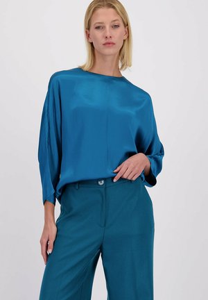 Vrouw in een losse, glanzende blauwe blouse en bijpassende blauwe hoge taillebroek voor een effen achtergrond.